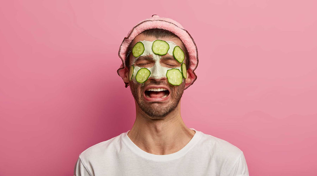 A man in a white shirt with a pink headband on and a green facemask with cucumbers across the upper half of his face, he has a pained expression on his face.