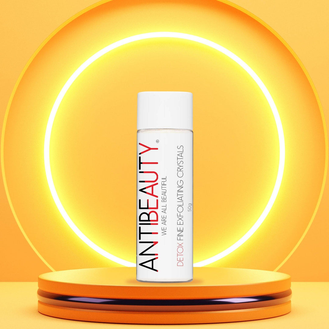 AntiBeauty Detox Fine Exfoliating Crystals - a gentle exfoliator for all skin types - on an orange podium in front of a glowing yellow background.