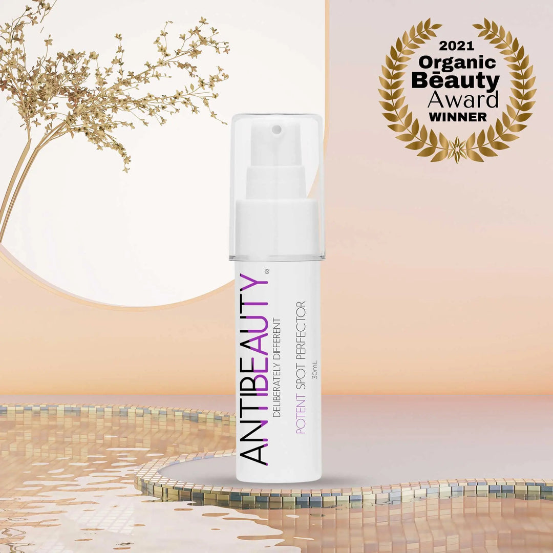 AntiBeauty Potent Spot Perfector - a powerful acne controlling spot treatment - on a pastel platform near water with a plant behind it.