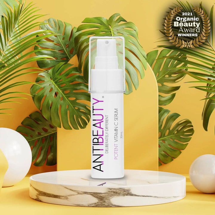 AntiBeauty Potent Vitamin C Serum - a powerful brightening and refreshing serum - on a white marble podium in front of a yellow background with plants.