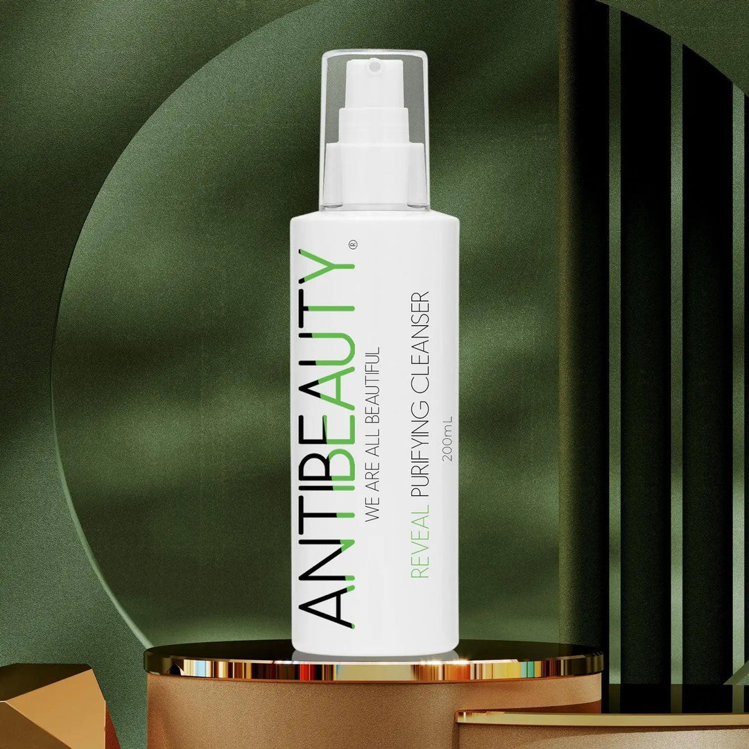 AntiBeauty Reveal Purifying Cleanser - a gentle and effective solution for oily or acne prone skin - on a gold platform in front of a green background.