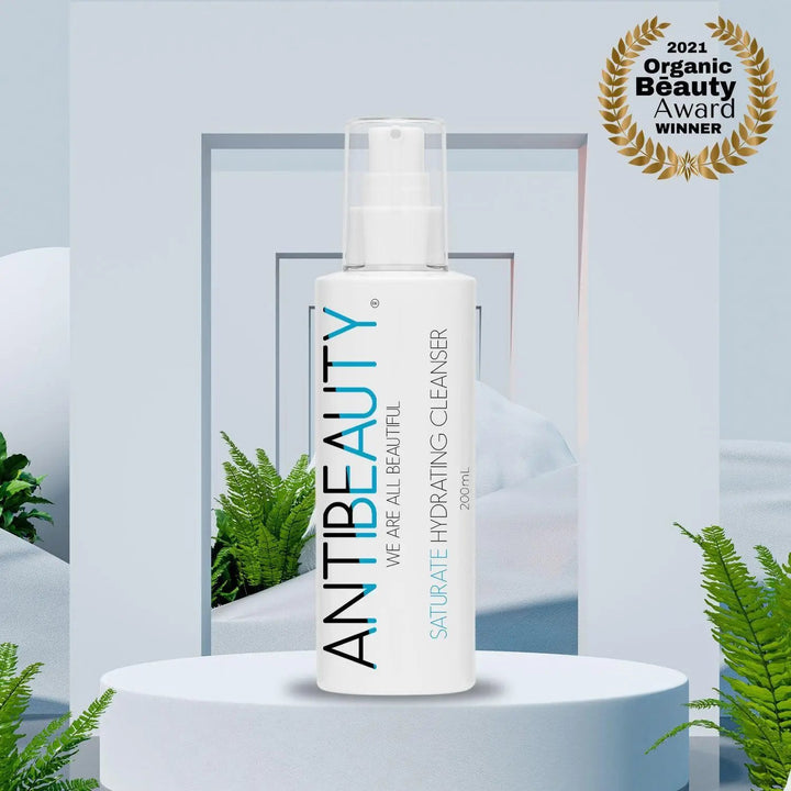 AntiBeauty Saturate Hydrating Cleanser - a gentle and effective solution for normal or dry skin - on a pastel blue platform background with plants. Award Logo Visible